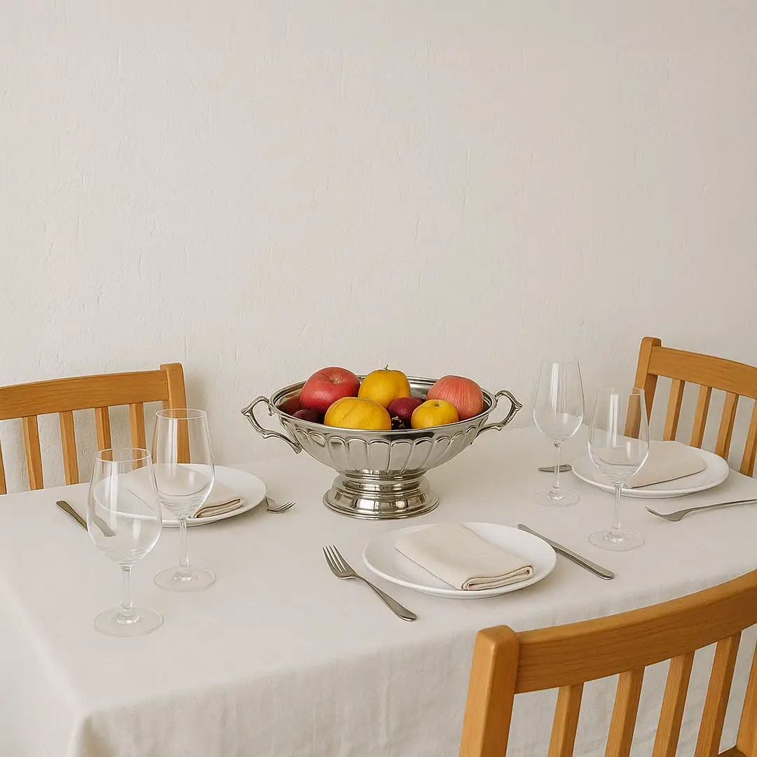 A silver fruit bowl filled with fresh apples and lemons placed at the center of a neatly set dining table with wooden chairs creating a warm home dining atmosphere.