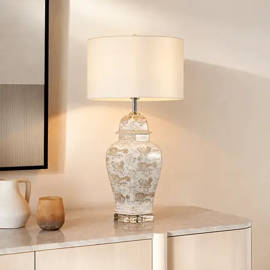 A table lamp with a white drum shade and a decorative ceramic jar base featuring a gold-toned floral pattern, placed on a light stone console table in a warm interior setting.